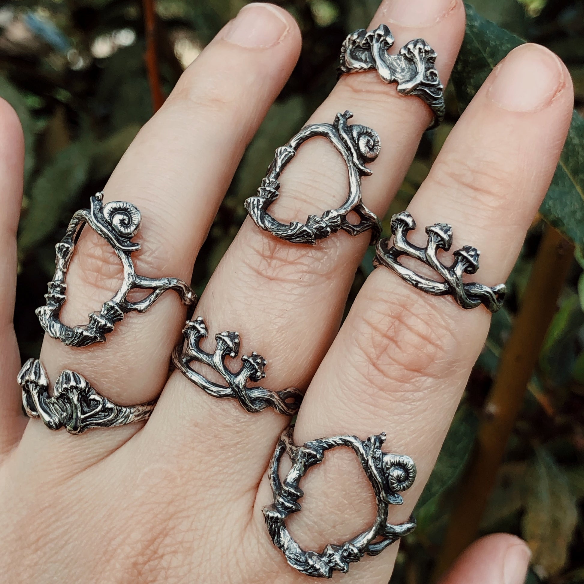 Hand wearing multiple oxidized silver rings with botanical sculptural designs, forest background