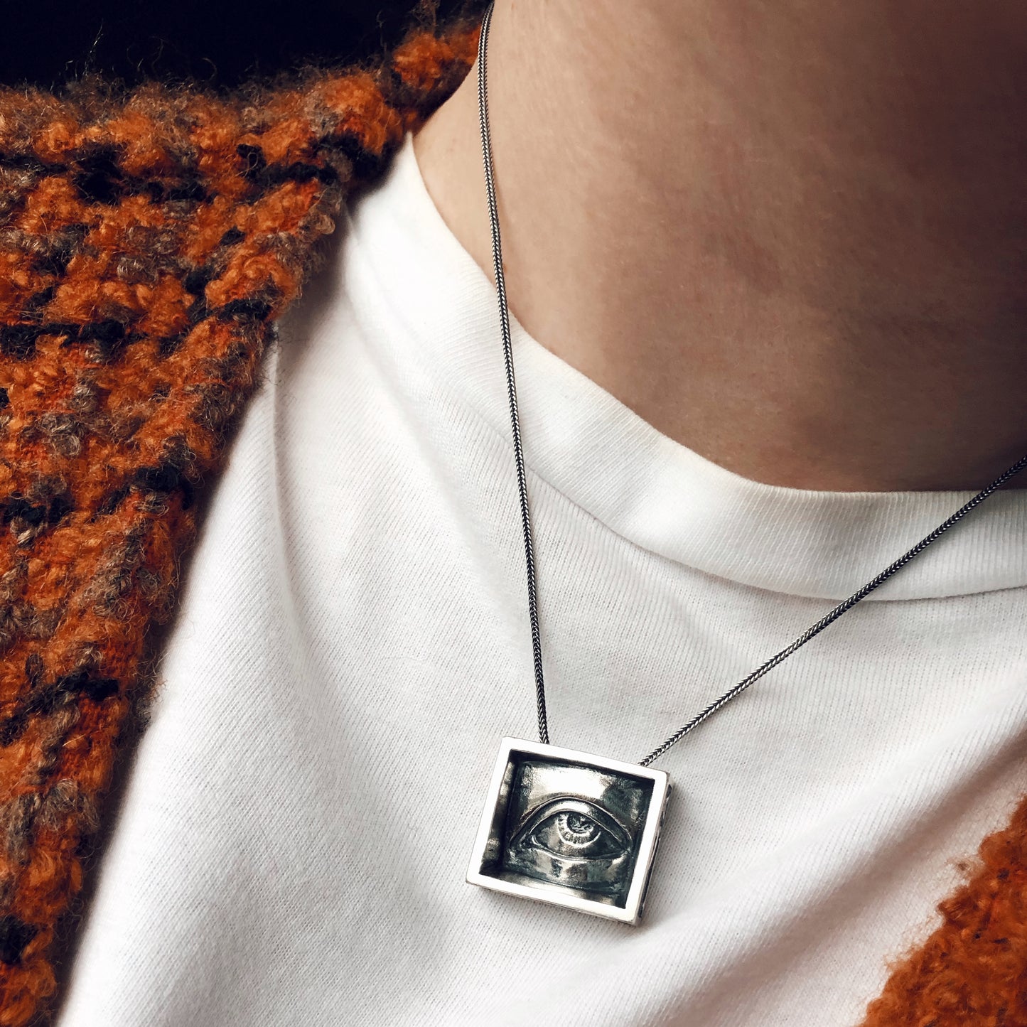 Sculptural silver eye pendant worn on a black chain over a white shirt, framed by a rust-toned wool cardigan. Gothic aesthetic with handcrafted detail.
