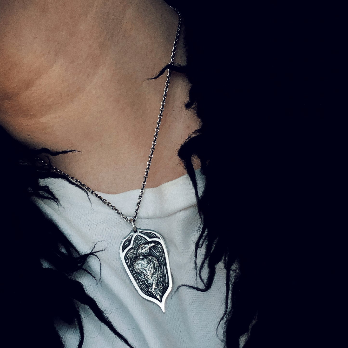 Raven pendant in oxidized sterling silver worn over a white shirt, framed by black fabric, sculptural and gothic in style