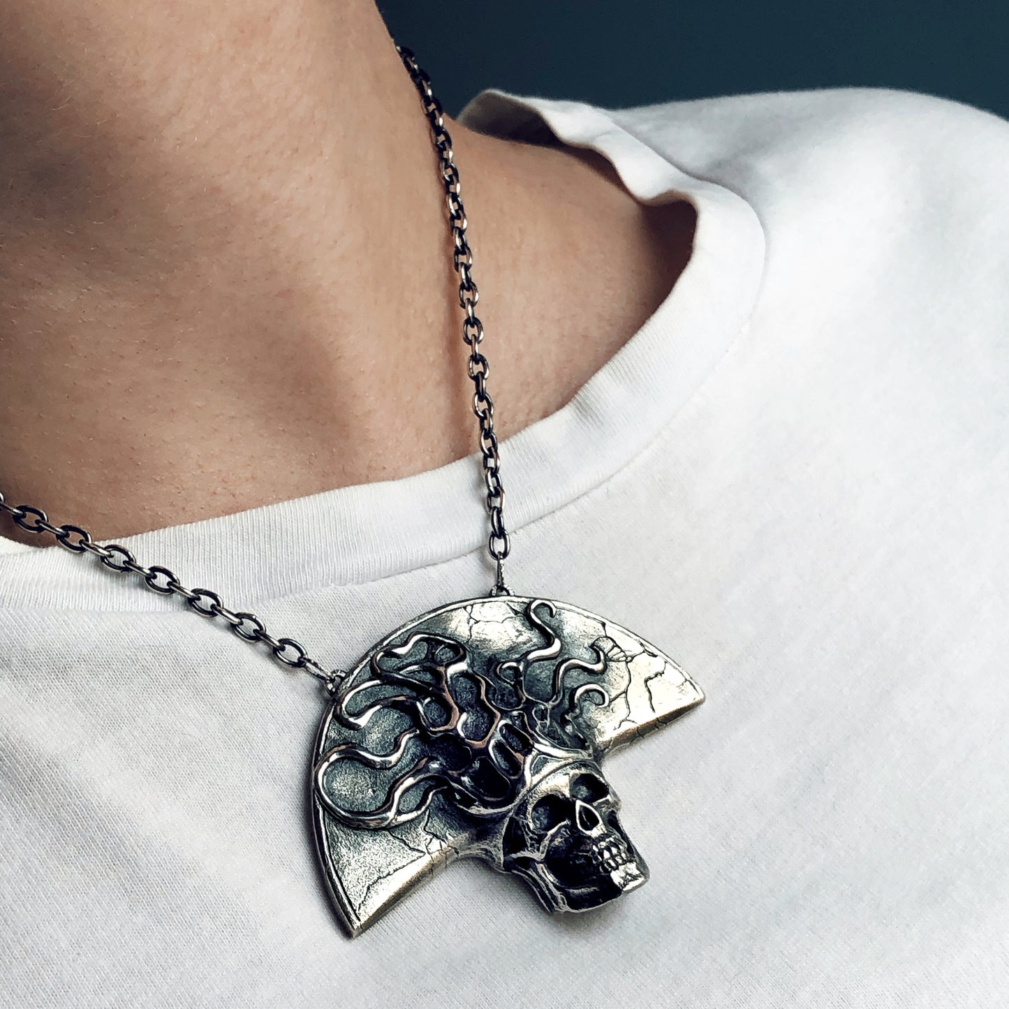 Crowned skull talisman in oxidized sterling silver worn on chest, sculptural and textured with root-like details, suspended from a thick silver chain over a white shirt