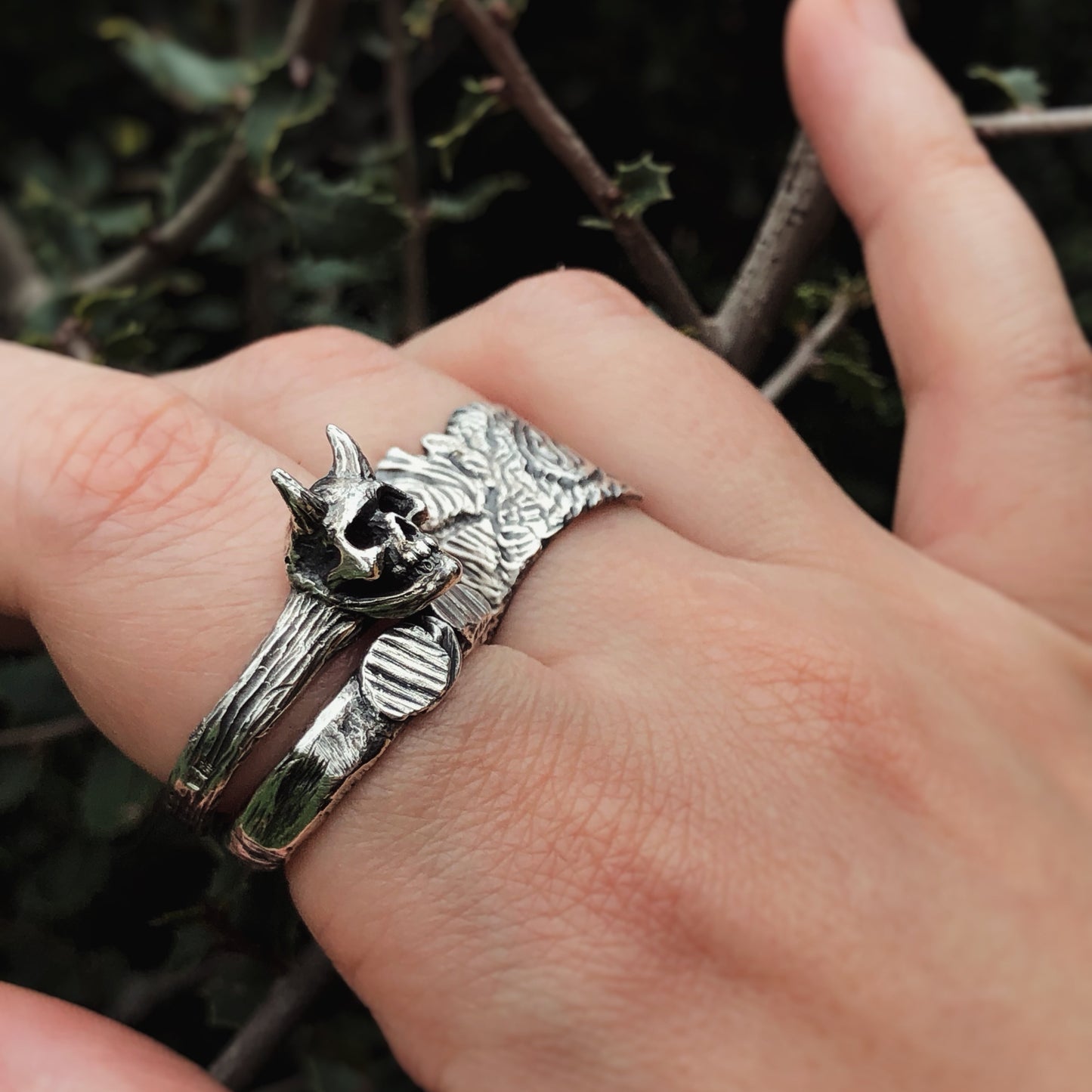 Sterling silver ring with horned skull design worn on a hand, photographed against thorny forest branches. Gothic artisan jewelry with raw, forest-inspired texture and dark woodland aesthetic.