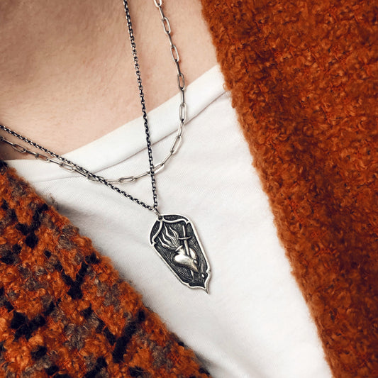 Sterling silver sacred heart pendant with sword, worn layered with silver chains. Victorian-inspired, gothic jewelry with religious symbolism.