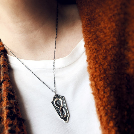 Ouroboros serpent pendant in oxidized sterling silver, worn over a white shirt and rust-coloured coat, sculptural and symbolic in form