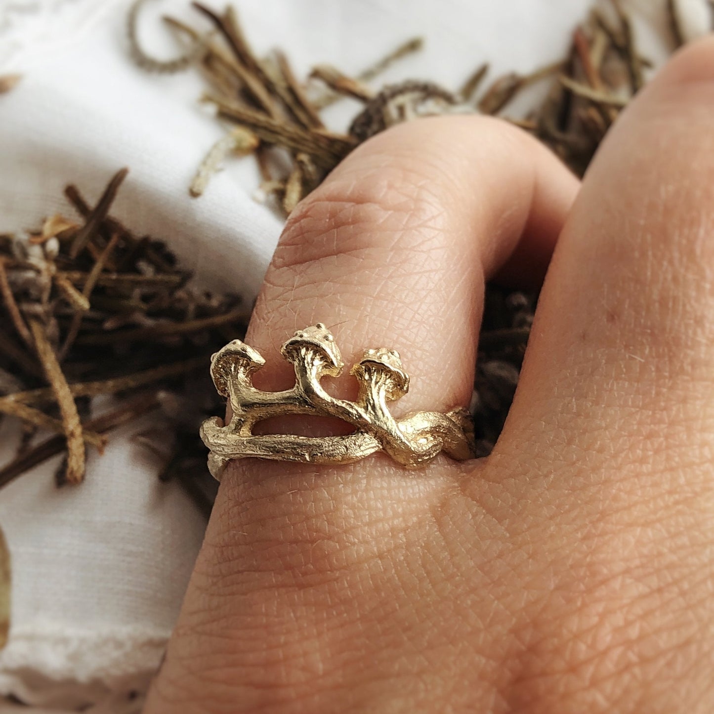 Close-up of a hand wearing a sculpted 9k gold ring with three tiny mushrooms rising from textured branches. The ring has an organic, woodland design and a soft matte finish. Styled on a natural linen cloth with dried forest twigs in the background. Nature-inspired gold ring, fairycore jewelry, handmade botanical talisman.