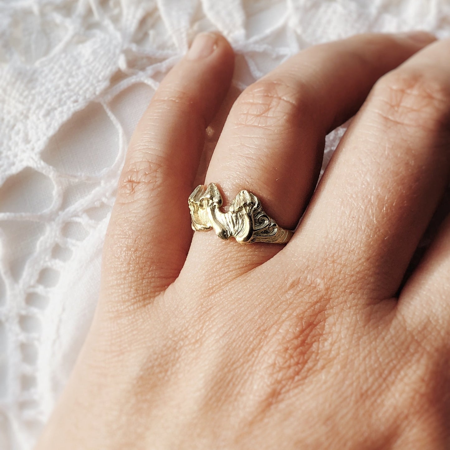 Gold ring with sculpted mushrooms, worn on a hand resting on white lace. Botanical gold jewelry, symbolic talisman ring, nature-inspired design.