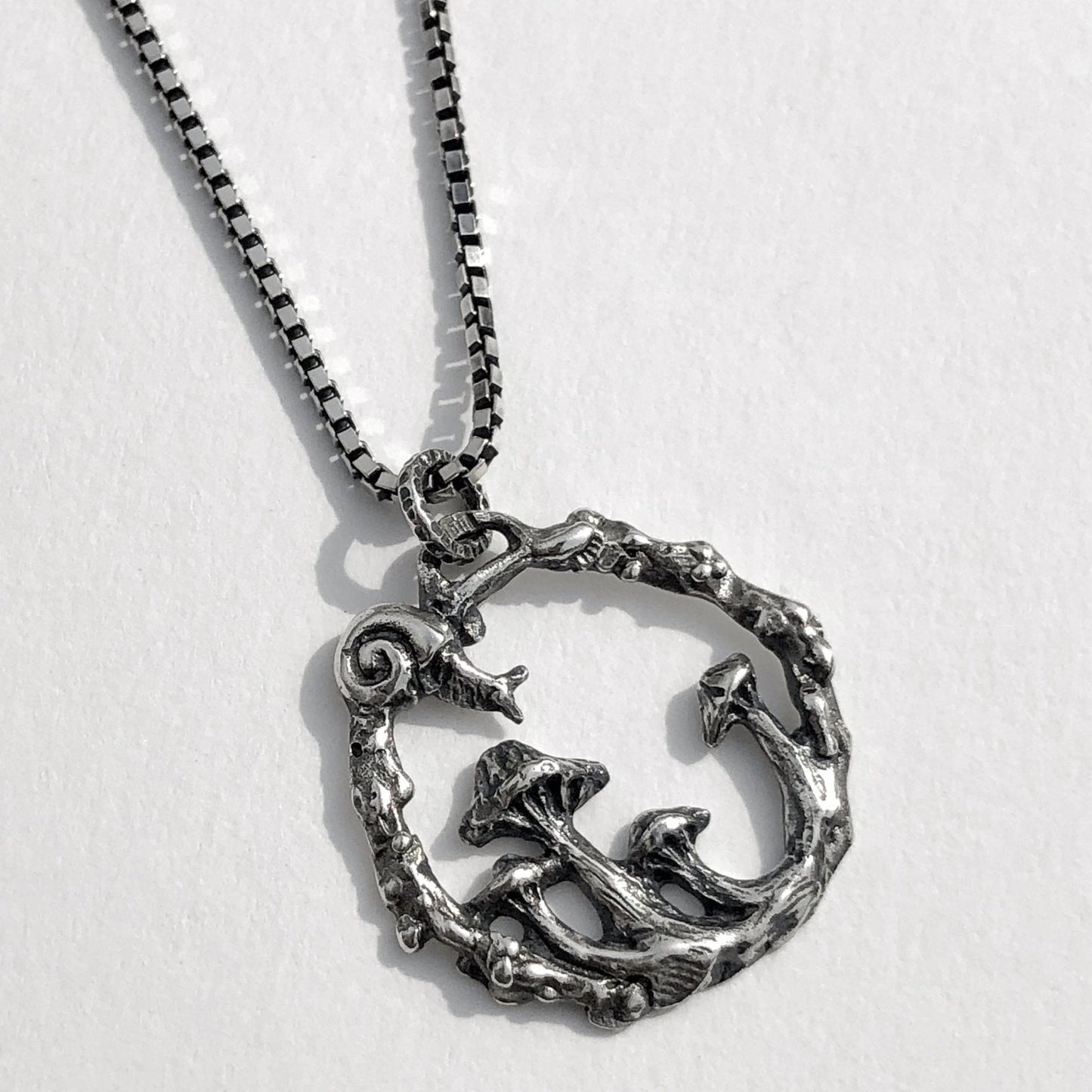 Sterling silver pendant featuring a sculpted snail and cluster of wild mushrooms inside a textured circular frame, inspired by enchanted forest scenes. White background.