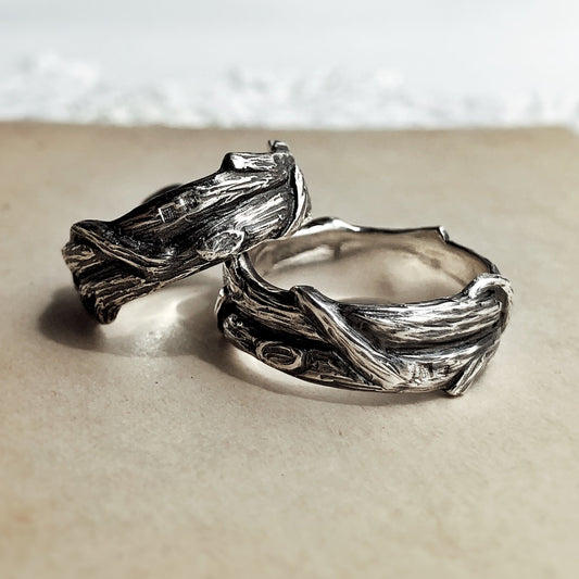 Two sterling silver rings with bark-like texture and entwined branches, placed side by side on a beige surface. Handcrafted with an oxidized finish and organic shape, these nature-inspired silver bands are designed as symbolic rings for soulmates, alternative wedding rings, or botanical jewelry lovers drawn to forest symbolism and artisan design.